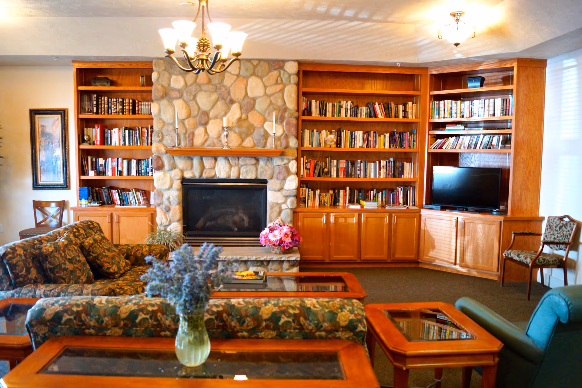 A cozy living room with floral patterned sofas and wooden coffee tables with glass tops. The room features a stone fireplace in the center with candles on the mantel, flanked by wooden bookshelves filled with books. A flat-screen TV is placed on the right bookshelf. There are two chairs, one on each side of the room, and a vase with purple flowers on the coffee table in the foreground. The room is warmly lit by ceiling lights.
