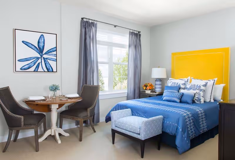 A bright bedroom with a large window covered by gray curtains. The room features a bed with a yellow headboard, blue bedding, and multiple decorative pillows. At the foot of the bed is a small upholstered bench. To the left, there is a small round wooden table set for two with dark upholstered chairs. A framed artwork of a blue leaf hangs on the wall above the table. A nightstand with a lamp and a flower arrangement is beside the bed.