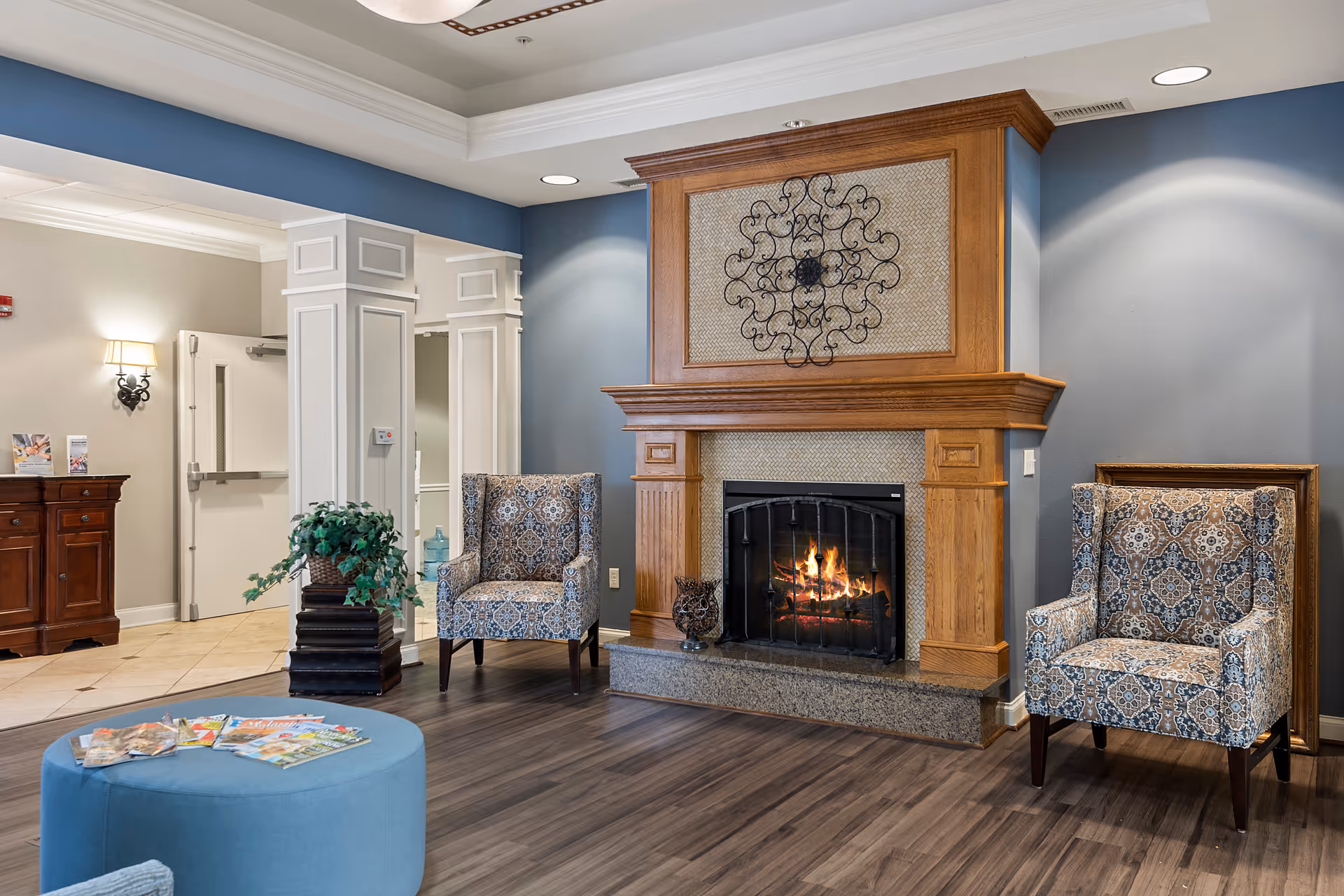 A cozy interior lounge area featuring a lit fireplace with a wooden mantel and decorative metal wall art above it. Two patterned armchairs are positioned on either side of the fireplace. In the foreground, there is a round blue ottoman with magazines on top. The room has wood flooring and soft lighting, with a small wooden cabinet and a plant in the background.