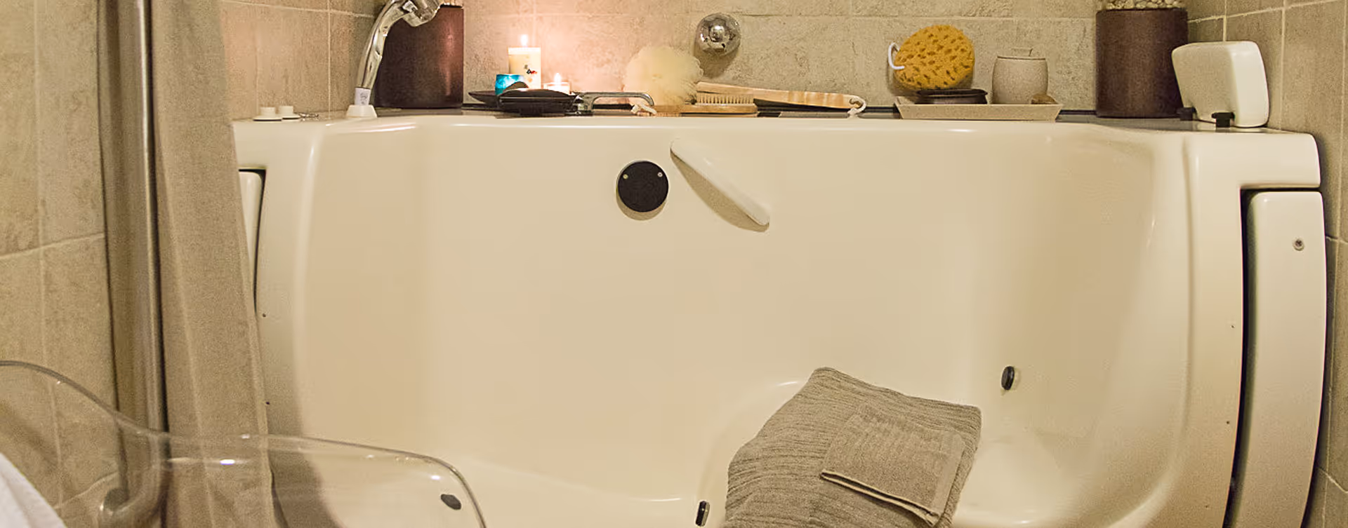 A white walk-in bathtub with a folded towel inside. On the edge of the tub, there are various bath accessories including brushes, a sponge, candles, and containers. The surrounding walls are tiled in a beige color.