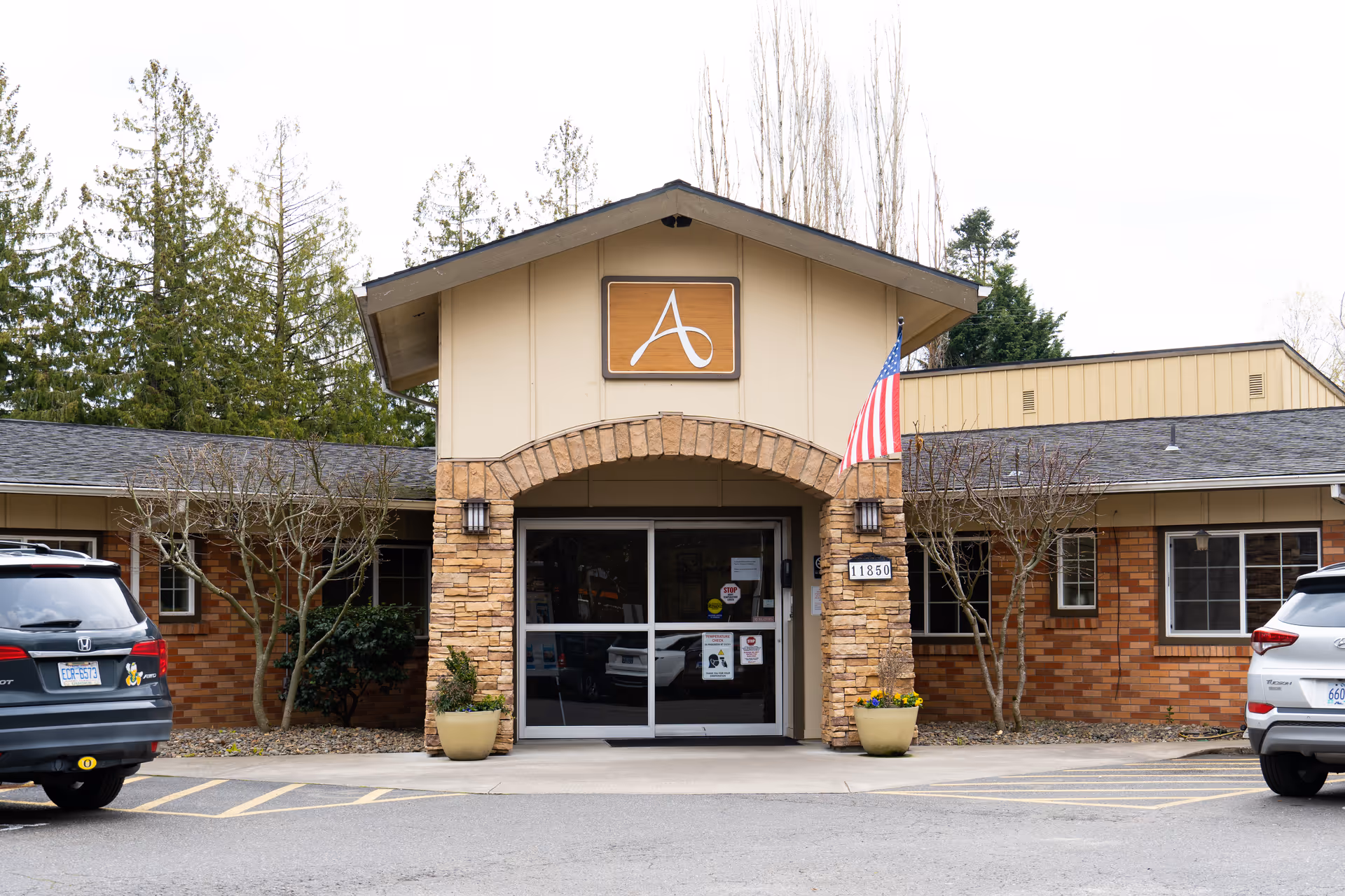 Front entrance of a single-story brick and stone senior living facility with an arched entryway, a mounted 'A' logo, an American flag, and parked cars.