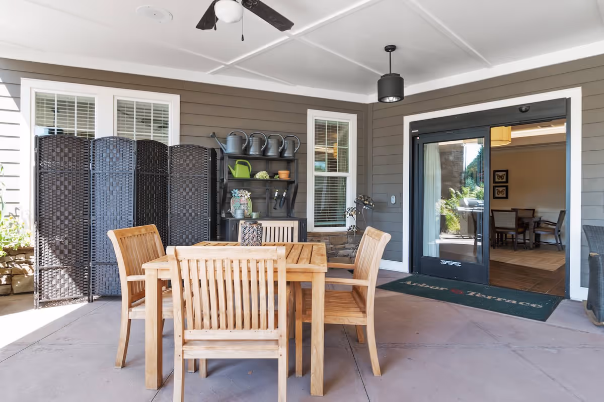 Covered outdoor patio area with a wooden table and four wooden chairs. There is a black ceiling fan and a black hanging light fixture. Against the wall are two windows with white blinds, a black folding screen, and a black shelving unit holding several watering cans and gardening supplies. A glass door leads inside to a dining area. A green doormat with the text 'Arbor Terrace' is placed in front of the door.