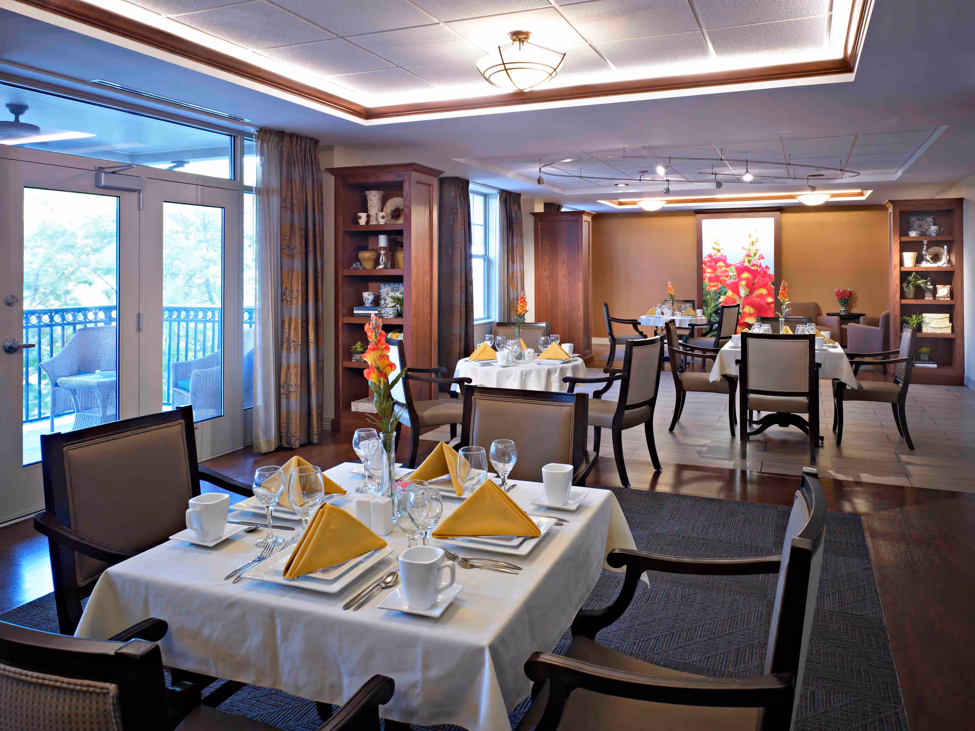 A well-lit dining room with several tables set for a meal, each table covered with white tablecloths, yellow folded napkins, glassware, and white coffee cups. The room features wooden chairs with cushioned seats, decorative shelves with vases and plants, large windows with curtains, and a door leading to an outdoor balcony with seating. The ceiling has recessed lighting and a modern light fixture.