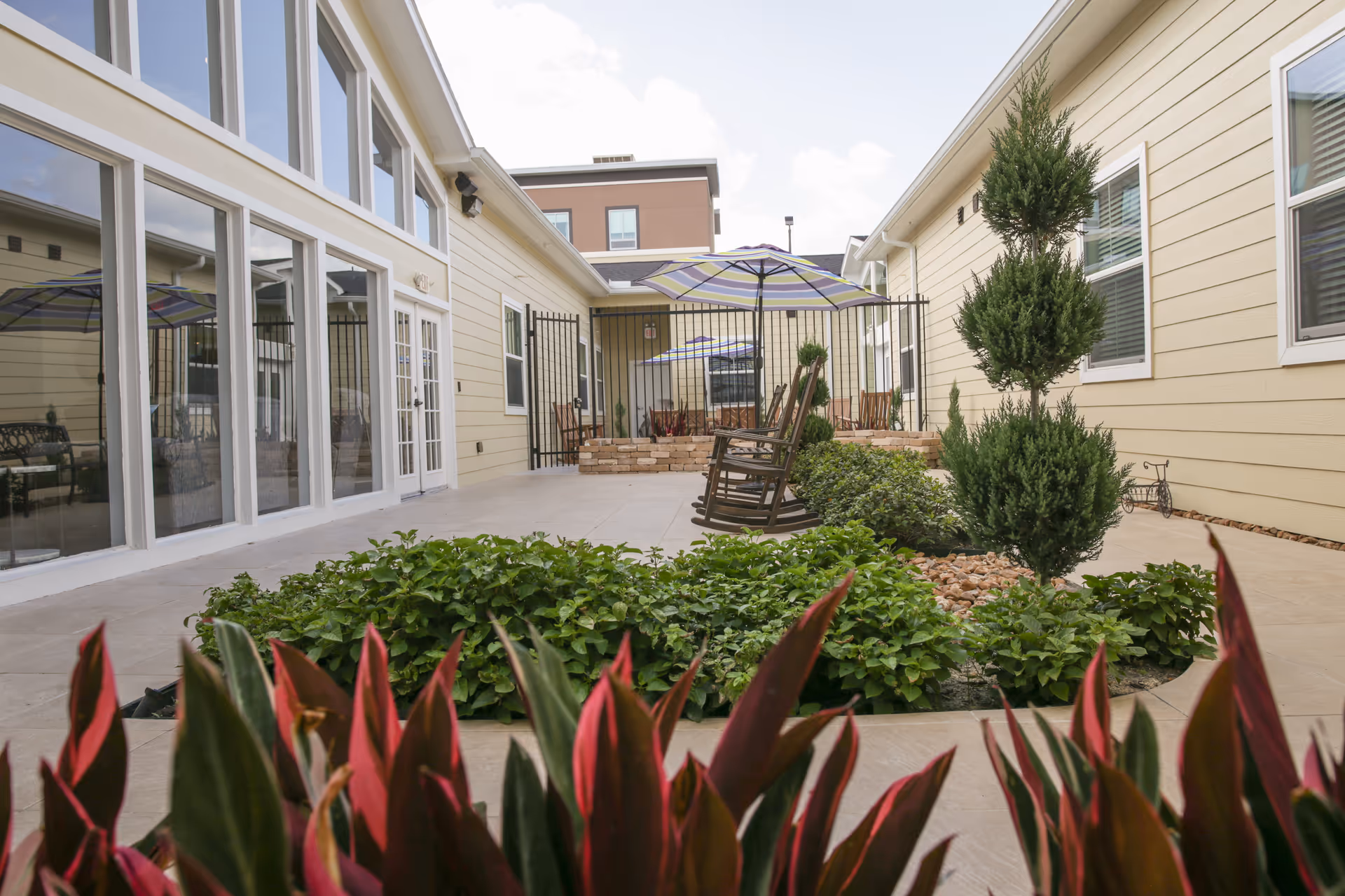 Outdoor courtyard area with beige siding buildings on both sides, a tiled floor, green plants and shrubs in the center, two wooden rocking chairs, and striped umbrellas providing shade.