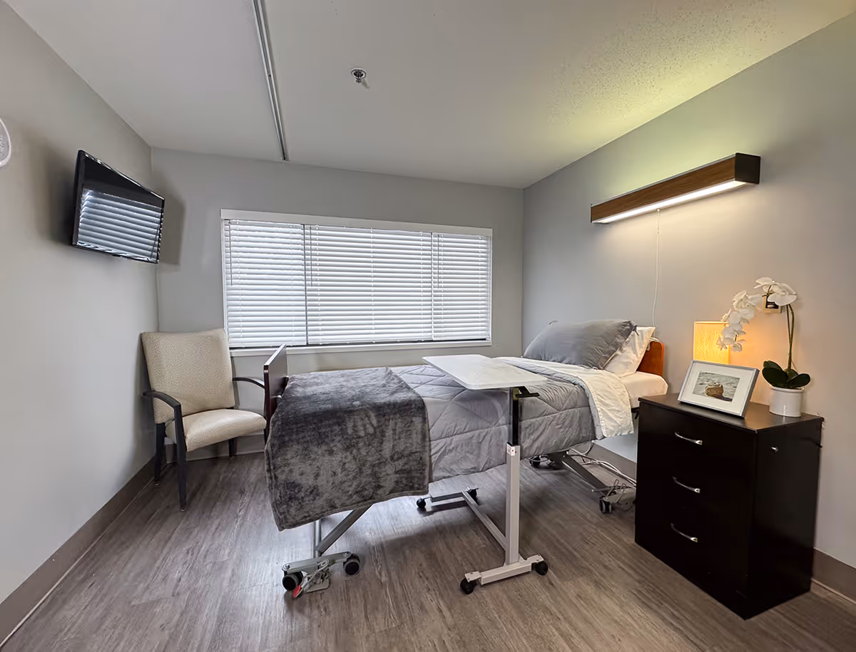A clean and simple nursing facility room with a hospital bed covered in gray bedding and a small overbed table. There is a beige armchair next to the window with closed blinds, a wall-mounted TV, and a dark wooden nightstand with a lamp, a framed photo, and a white orchid plant. The room has light gray walls and wood-look flooring.