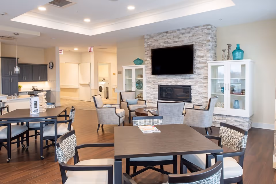 A modern senior living common area featuring multiple tables and chairs with cushioned seats, a stone fireplace with a mounted flat-screen TV above it, and two white display cabinets with decorative vases. The room has wood flooring, recessed lighting, and an open view into a kitchen area with dark cabinets and a white refrigerator.