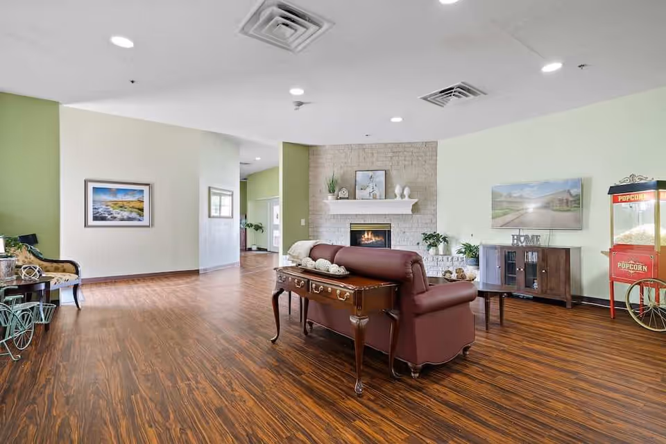 A spacious living room area with wooden flooring, a brown leather sofa facing a white brick fireplace with decorative items on the mantel. There is a TV mounted on the wall above a wooden cabinet, a popcorn machine to the right, and a framed picture on the left wall. The room has light green and white walls with recessed ceiling lights.