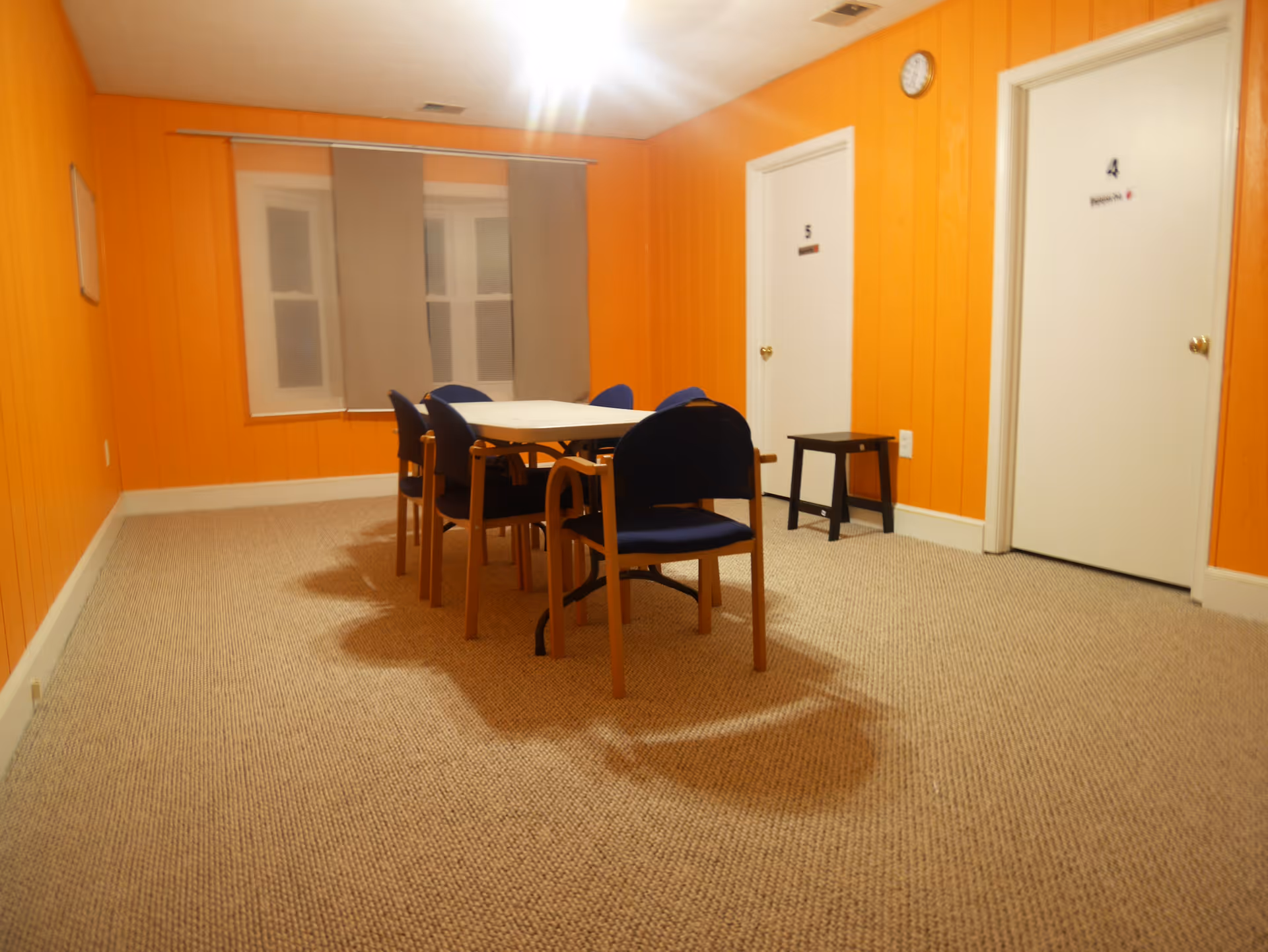 A small room with bright orange walls and beige carpet. There is a rectangular table in the center surrounded by six blue cushioned chairs with wooden frames. Two white doors labeled with numbers 4 and 5 are visible on the right wall, along with a small black stool. A window with beige blinds is on the far wall.