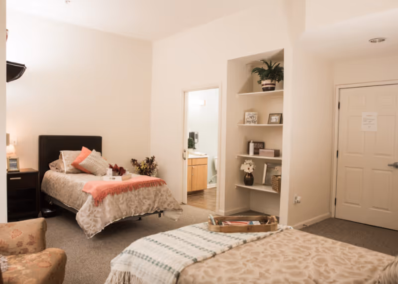 A cozy bedroom in a senior living facility with two beds, one with a coral throw blanket and decorative pillows, and the other with a green and white striped blanket. There is a floral armchair in the foreground, built-in shelves with decorative items, and a door leading to a bathroom with a sink and toilet visible.