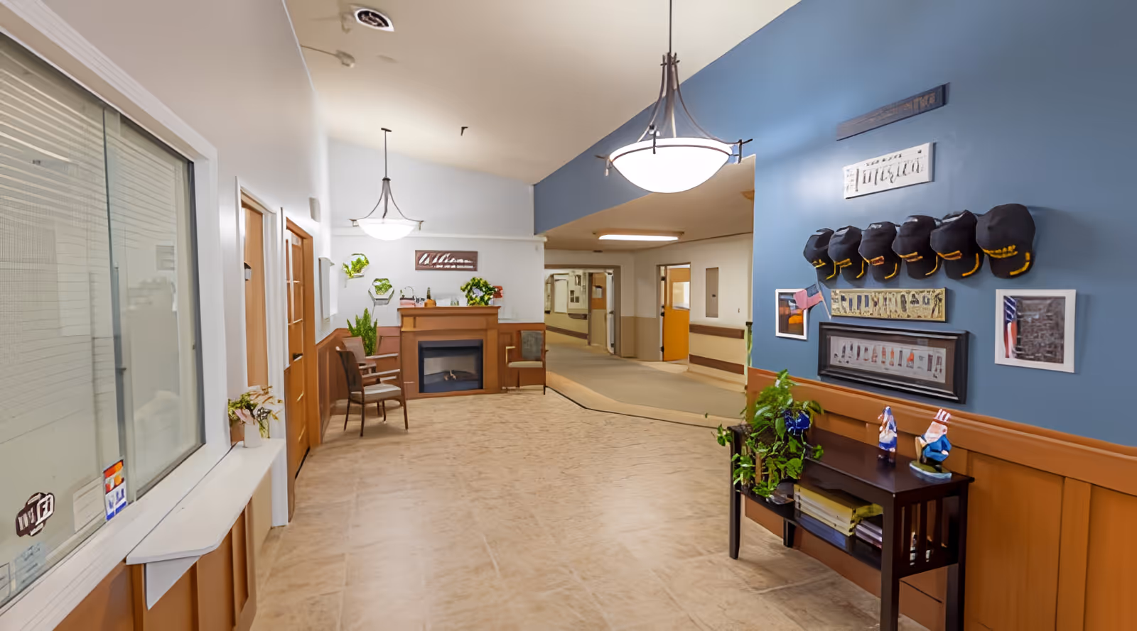 Interior hallway of Rose Haven Nursing Center featuring a seating area with chairs and a fireplace, decorative plants, wall art, and a display of hats on a blue accent wall. The hallway extends into other rooms with beige and white walls and overhead lighting.