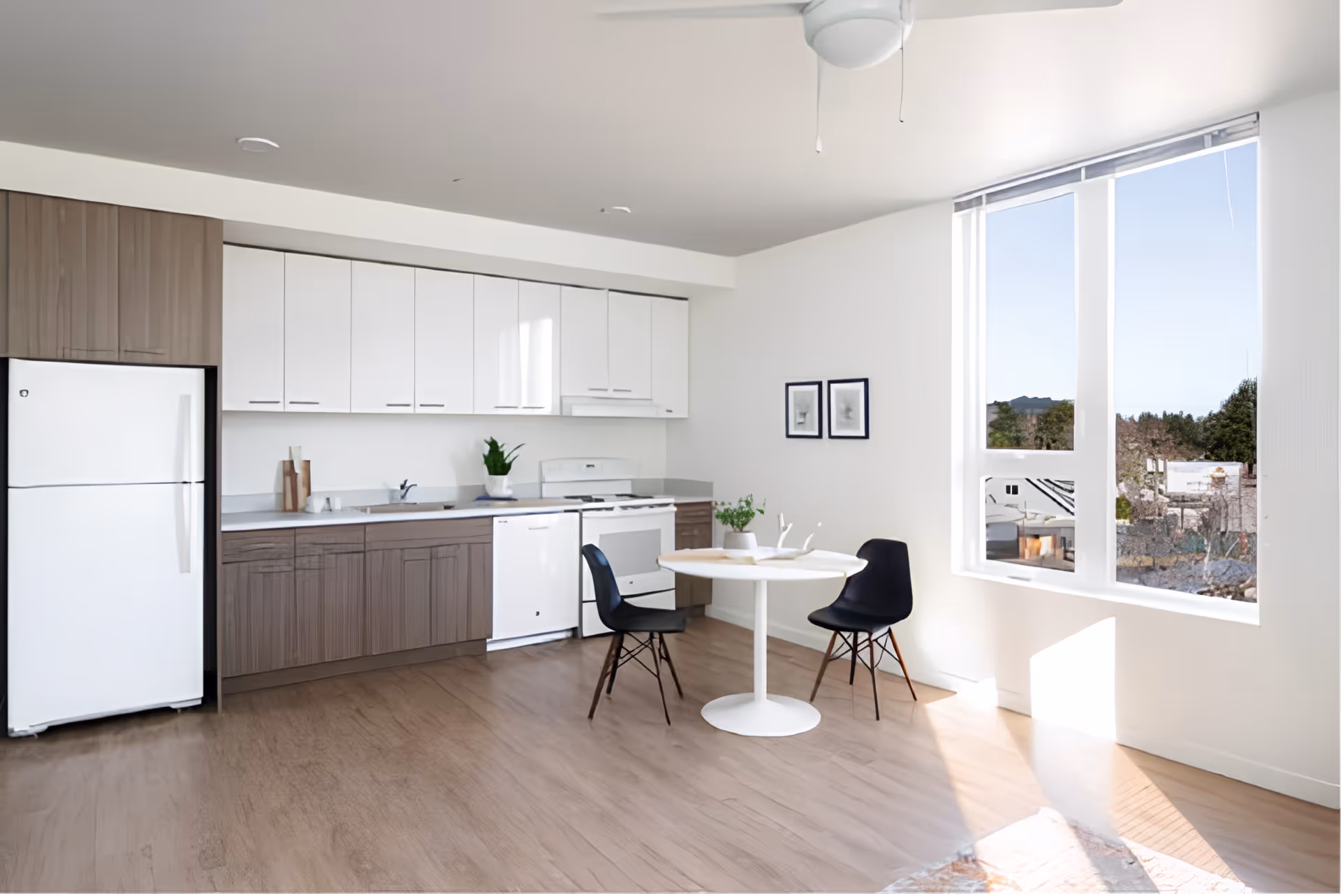 A bright kitchen area with white upper cabinets and wood-toned lower cabinets, a white refrigerator, stove, and dishwasher. There is a small round white dining table with two black chairs near a large window letting in natural light. The floor is wood, and two framed pictures hang on the wall.