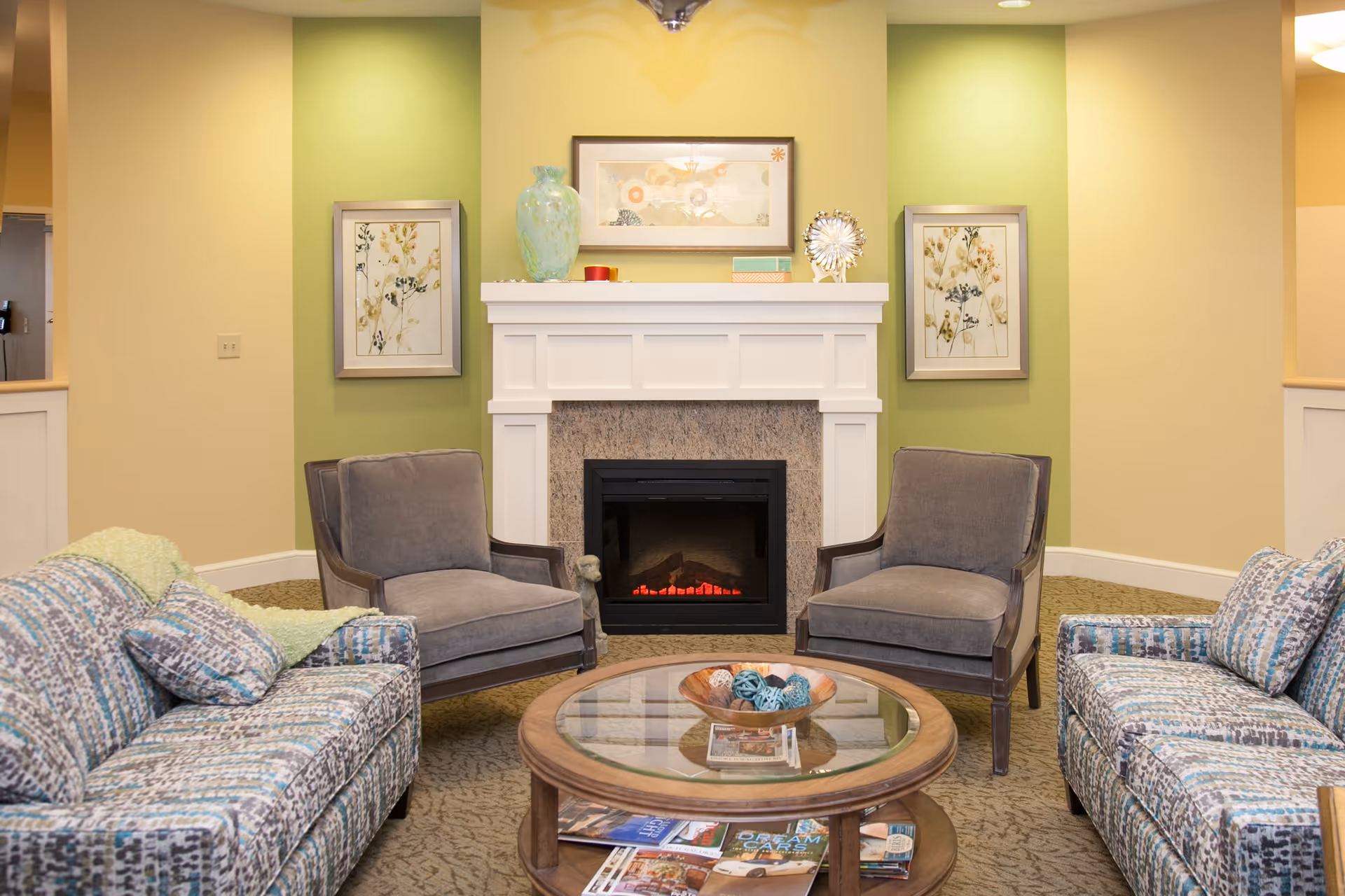 A cozy living room area with two patterned sofas and two gray armchairs arranged around a round glass coffee table. The table holds a decorative bowl with blue spheres and magazines on the lower shelf. Behind the seating is a white fireplace with a black insert, flanked by two framed floral artworks on a green accent wall. Additional decorative items are placed on the fireplace mantel.