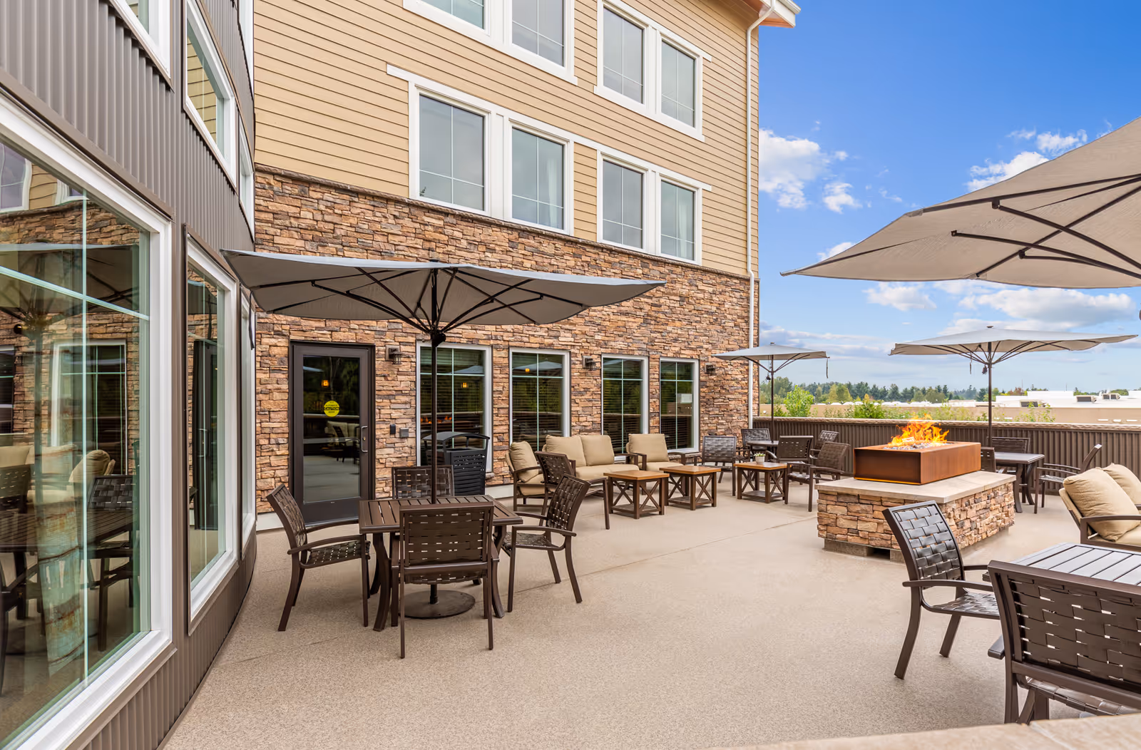 Outdoor patio area at Wesley Bradley Park featuring multiple seating arrangements with tables and chairs, large umbrellas for shade, cushioned sofas, and a central stone fire pit. The building exterior has stone and siding with several windows, and the sky is clear with some clouds.