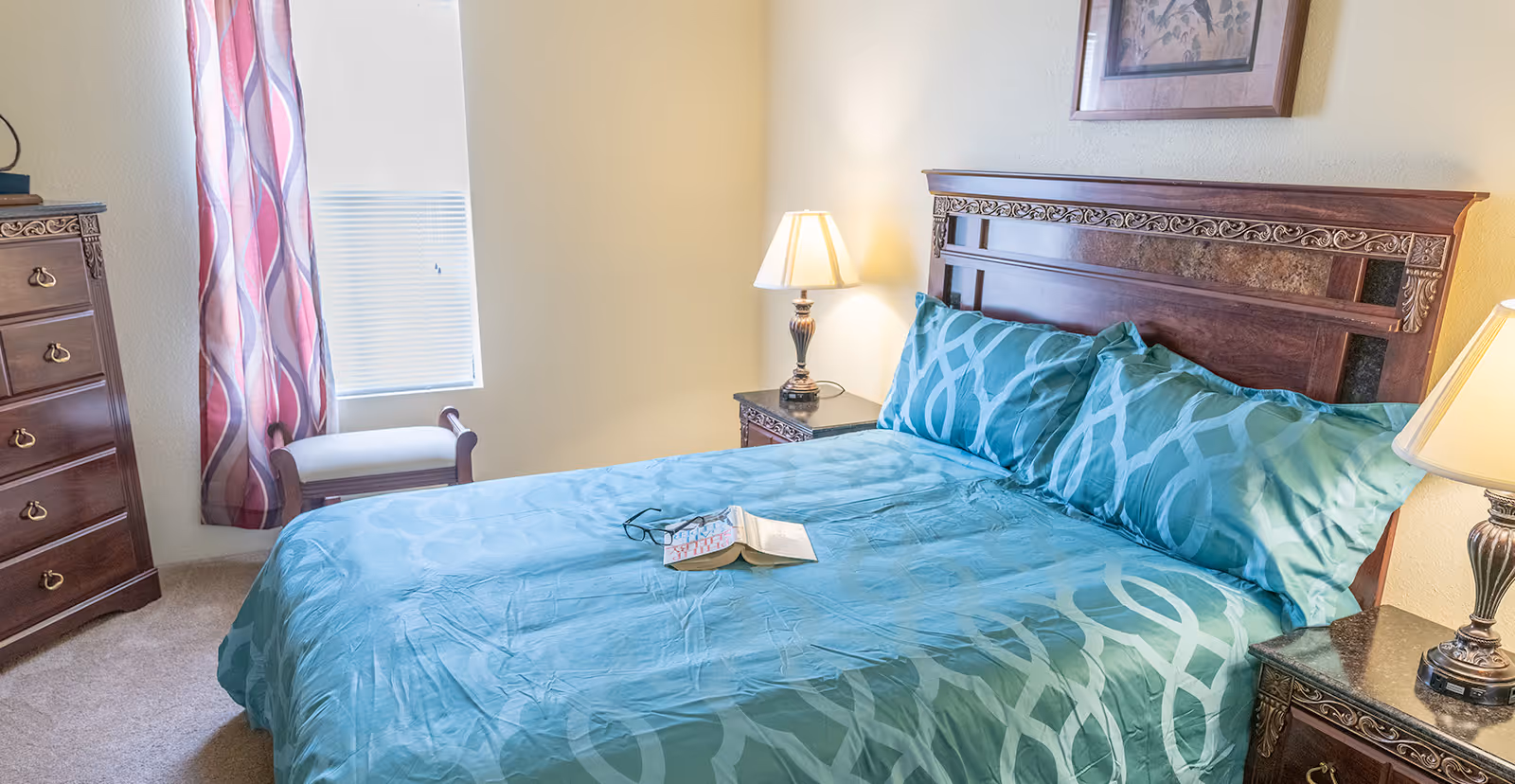 A bedroom with a large wooden bed featuring an ornate headboard, covered with teal bedding and pillows with a geometric pattern. On the bed lies an open book and a pair of glasses. There are two matching bedside tables with lamps on either side of the bed. A wooden dresser with brass handles is visible on the left side near a window with blinds and patterned curtains. A small wooden bench is placed under the window. A framed picture hangs on the wall above the bed.