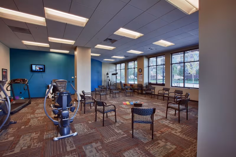 A spacious senior living exercise and activity room with several exercise machines including a treadmill and an elliptical. The room has multiple chairs arranged in a circle in the center, a blue accent wall with a mounted TV, large windows letting in natural light, and a patterned carpeted floor.