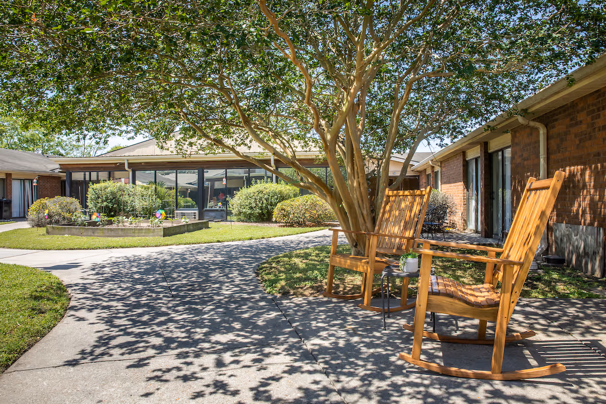 Outdoor courtyard area of a senior living facility with two wooden rocking chairs and a small table under a large tree, surrounded by a paved walkway, green grass, bushes, and brick buildings with large windows.