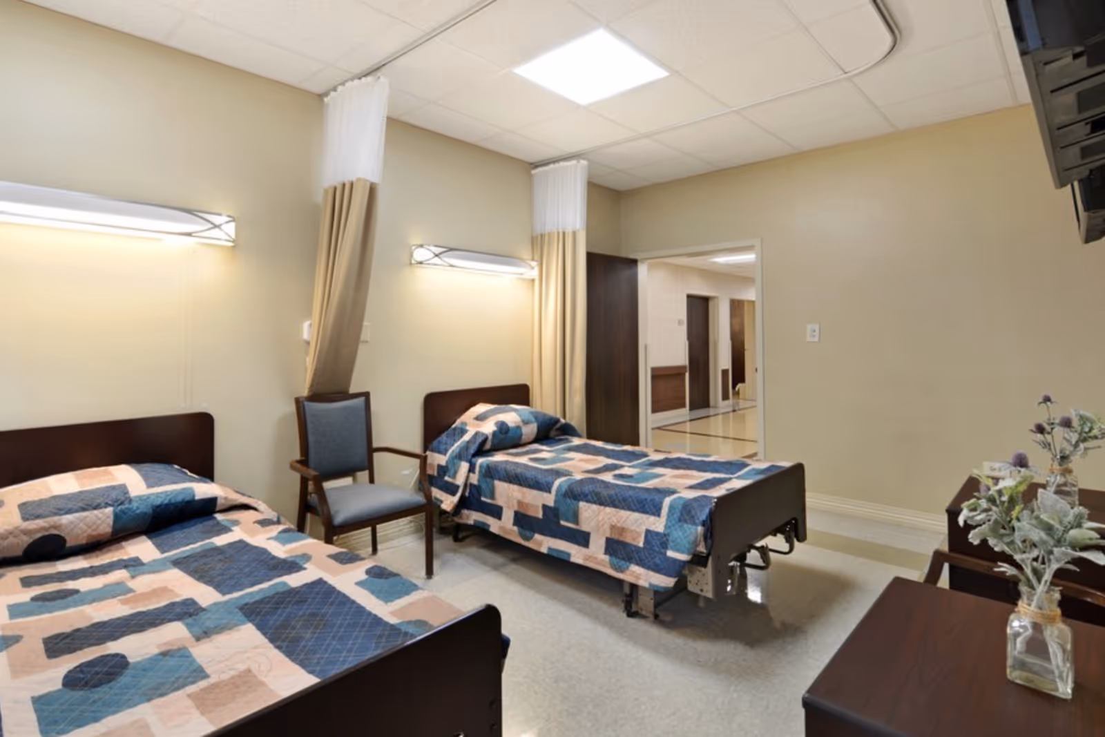 A room with two single beds covered with blue, beige, and brown patterned quilts. Between the beds is a wooden chair with a blue cushion. The room has beige walls, overhead fluorescent lighting, and a doorway leading to a hallway. There are two small tables with decorative plants on the right side of the image.