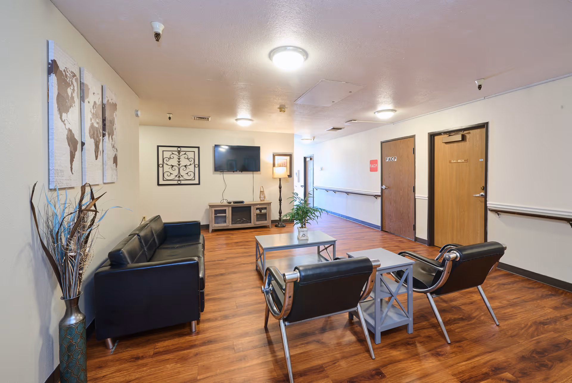 A well-lit common area with wooden flooring featuring a black leather sofa, two black chairs with metal frames, a coffee table with a plant, a TV mounted on the wall, and decorative wall art including a world map and a metal wall hanging. There are two wooden doors along the hallway with handrails on the walls.