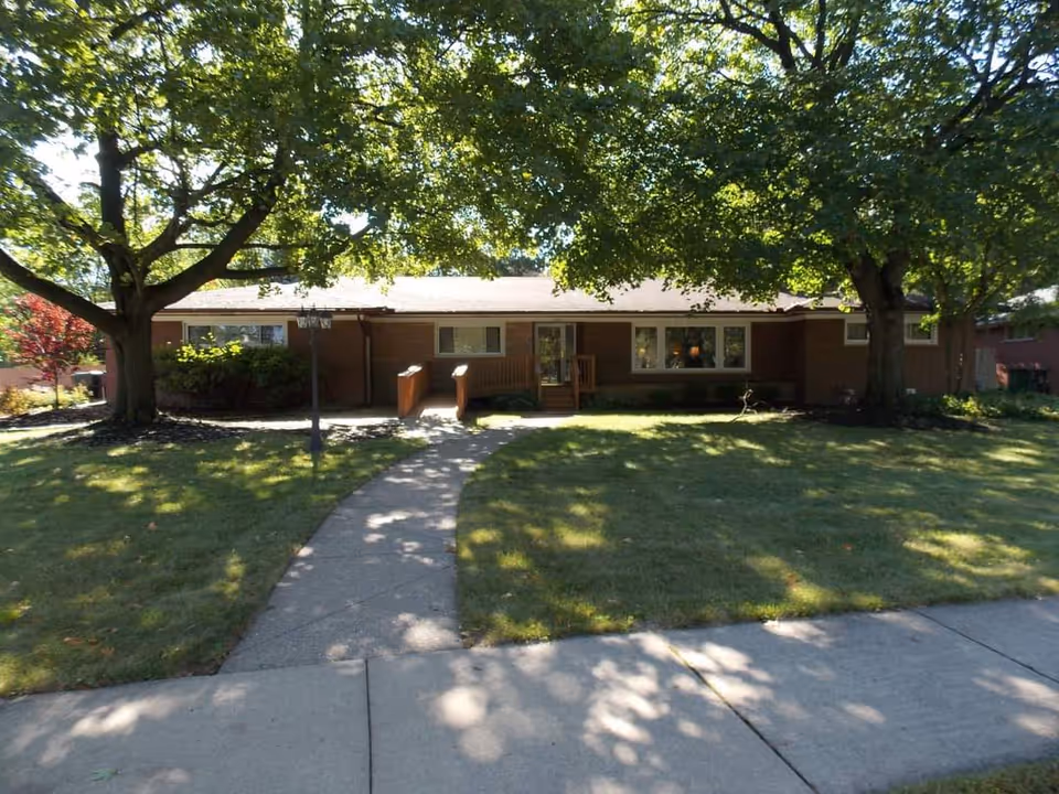 Front exterior view of a single-story senior living facility building with a brick facade, surrounded by large trees and a well-maintained lawn. A concrete pathway leads to the entrance, which has a small wooden ramp and steps.