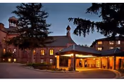 Exterior view of a senior living facility building at dusk with warm lights glowing from the windows. The building features a covered entrance with a circular driveway, surrounded by tall trees.