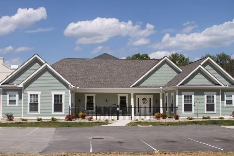 Single-story pastel green senior living building with a front porch and parking lot.
