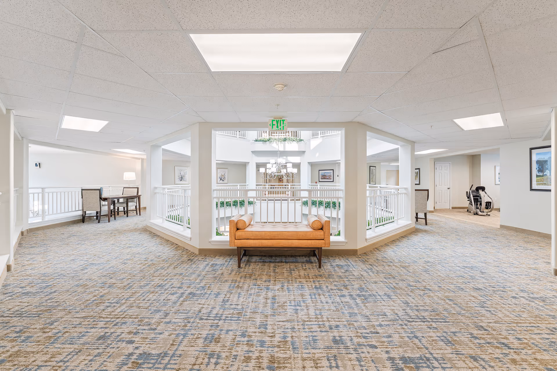 A bright and spacious interior hallway in a senior living facility with patterned carpet flooring, white walls, and a drop ceiling with fluorescent lighting. In the center, there is a cushioned bench with two bolster pillows placed against a railing overlooking a lower level. To the left, there is a small table with two chairs and a floor lamp. To the right, there is exercise equipment and framed artwork on the walls. The area appears clean and welcoming with natural light coming from the lower level.