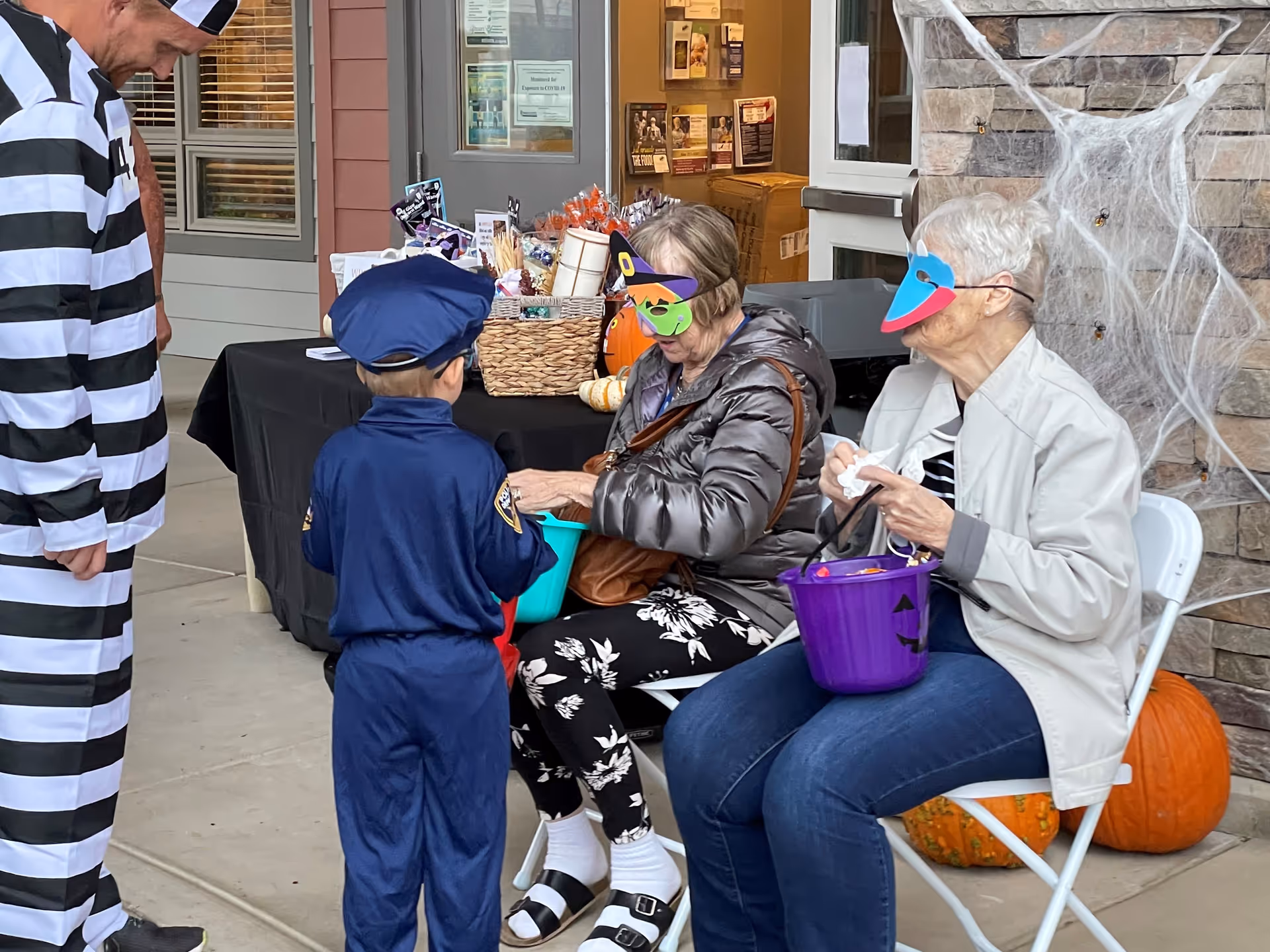 Two elderly women wearing colorful masks sit on white folding chairs outside a building decorated with Halloween pumpkins and spider webs. One woman is dressed in a puffy jacket and floral pants, holding a green bucket, while the other wears a light jacket and jeans, holding a purple bucket. A young boy dressed in a police officer costume and a man in a black and white striped prisoner costume stand nearby. A table covered with a black cloth and filled with Halloween treats is visible in the background.