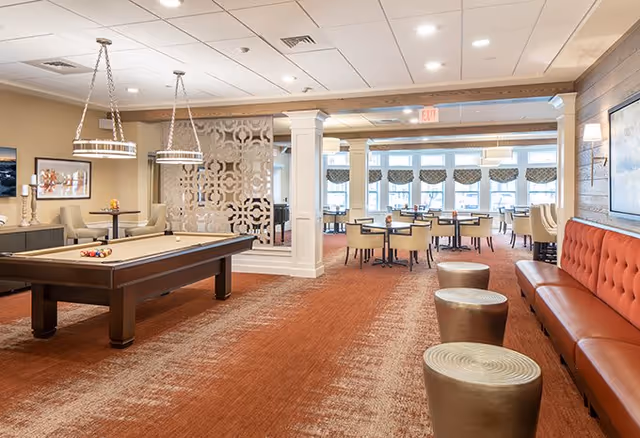 A spacious senior living common area featuring a pool table with balls arranged on it, modern pendant lights hanging from the ceiling, a decorative partition, several tables and chairs near large windows with patterned valances, and a long orange cushioned bench with round metallic stools along a wood-paneled wall.