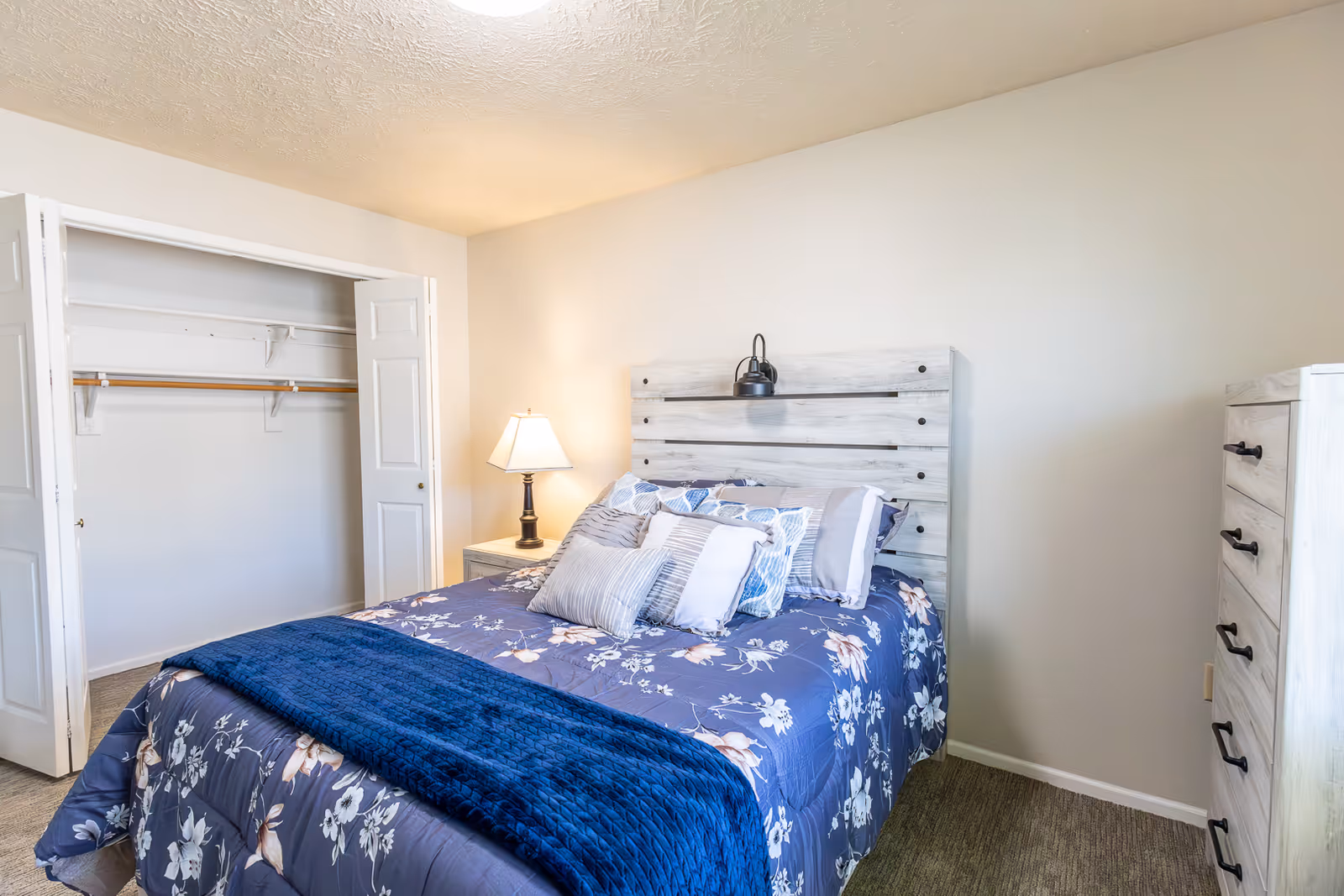 Well-lit bedroom with a bed dressed in blue floral bedding, a wooden headboard, a nightstand with a lamp, an open closet, and a dresser.