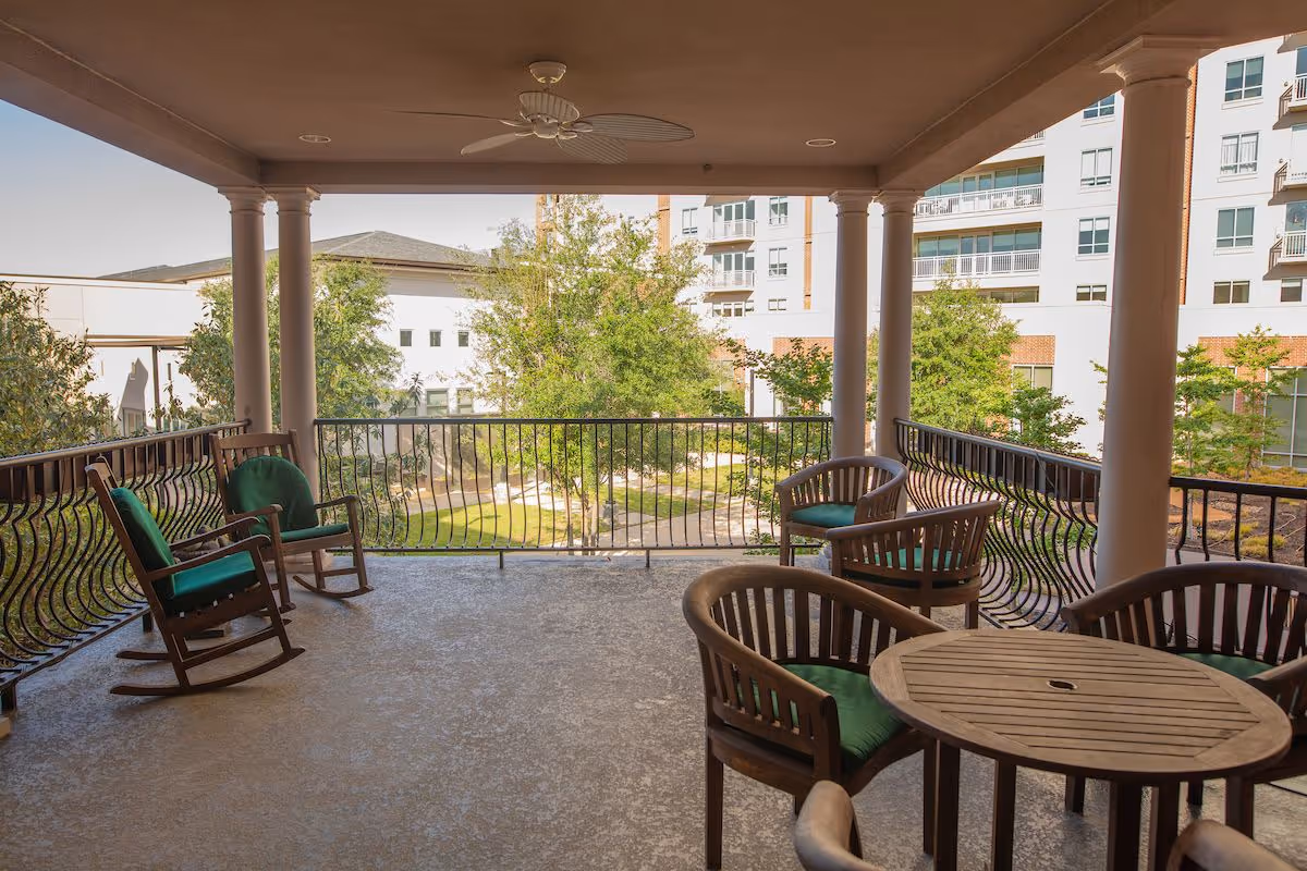 Covered outdoor patio area with wooden rocking chairs and cushioned chairs around a round wooden table, overlooking a landscaped garden and multi-story residential buildings in the background.