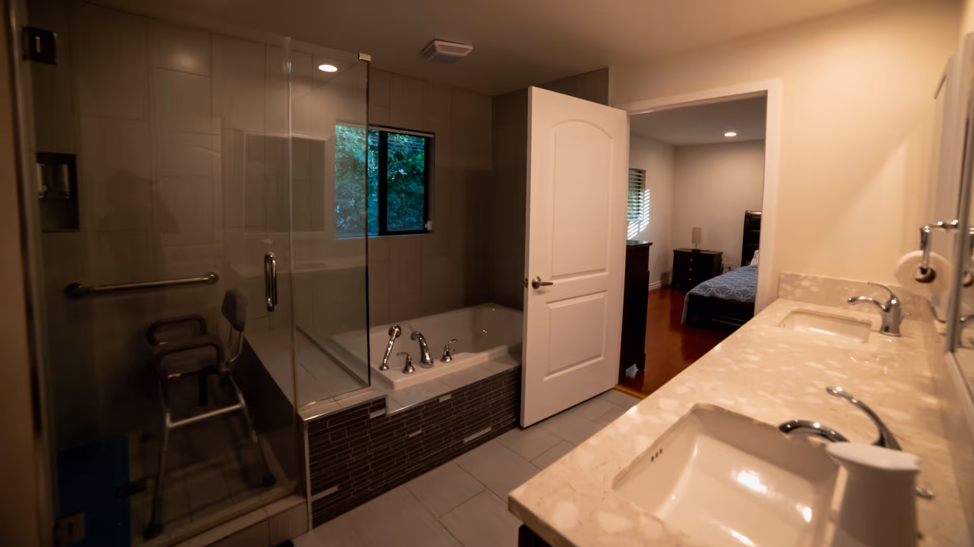A modern bathroom with a glass-enclosed shower featuring a shower chair, a bathtub with chrome fixtures, and a double sink vanity with a marble countertop. The bathroom door is open, revealing a bedroom with a bed and furniture in the background.