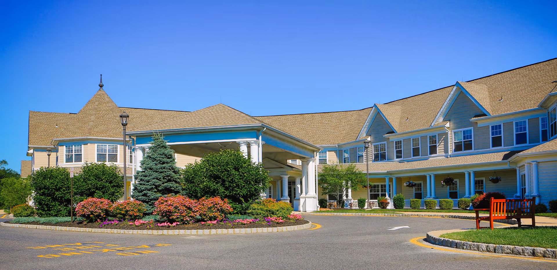 Exterior view of Brandywine Wall by Monarch facility showing a large building with beige siding, multiple windows, a covered entrance supported by white columns, landscaped bushes and flowers, a paved driveway, and a wooden bench on a grassy area under a clear blue sky.