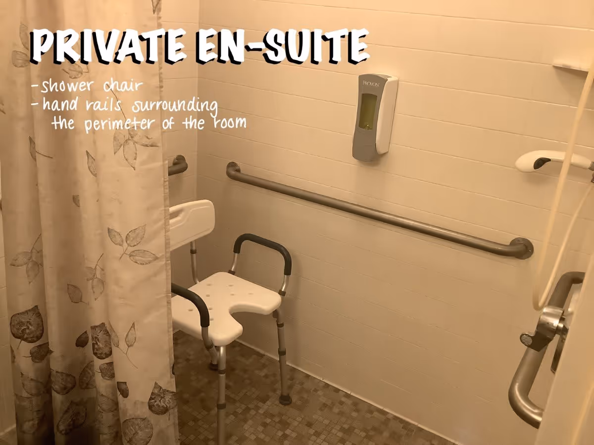 A private en-suite bathroom featuring a shower chair and hand rails surrounding the perimeter of the room. The walls are tiled in white, and there is a soap dispenser mounted on the wall. A shower curtain with a leaf pattern is partially visible.