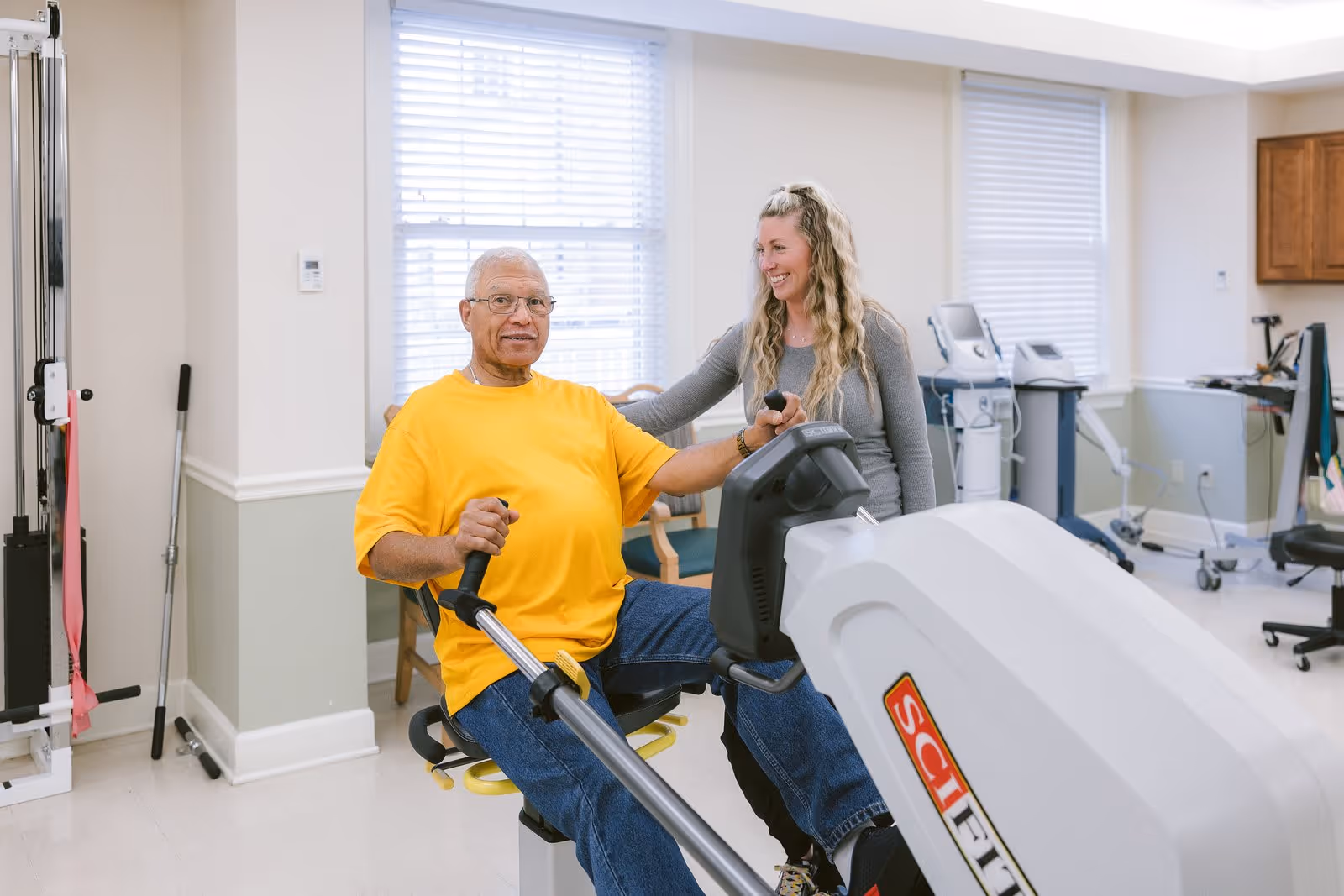 An elderly man in a yellow shirt is using a seated exercise machine in a rehabilitation or fitness room, assisted by a smiling woman with long blonde hair wearing a gray top. The room has exercise equipment, windows with blinds, and light-colored walls.