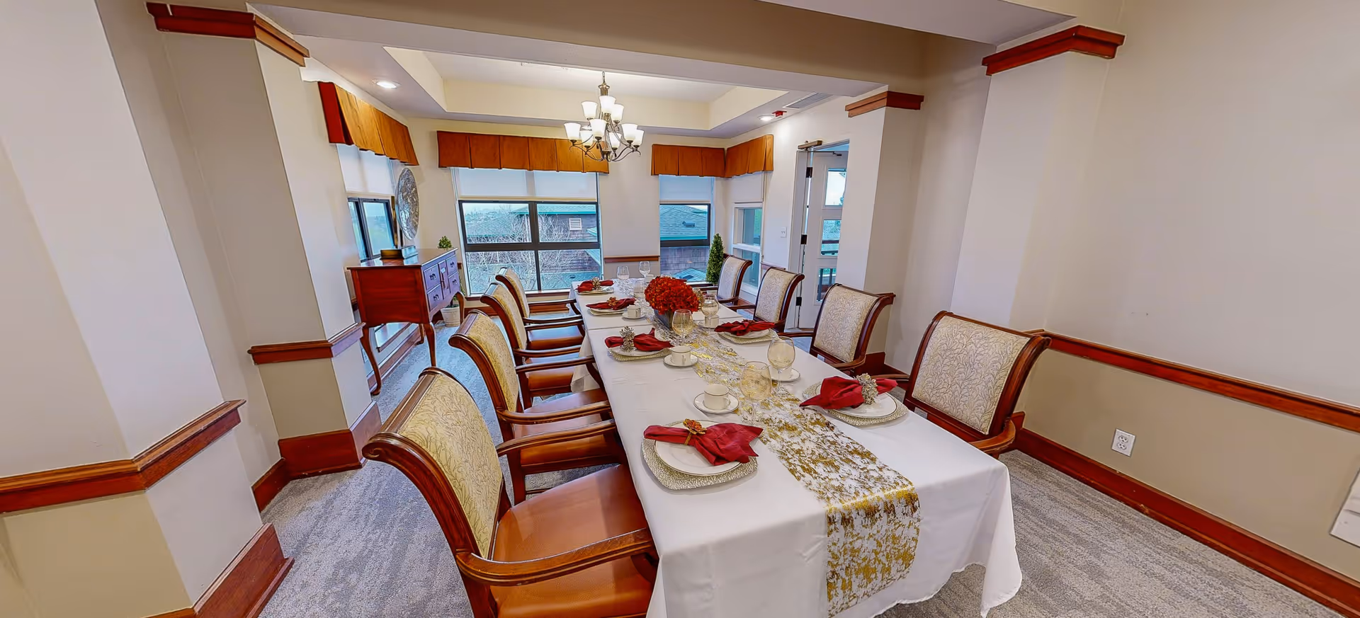 A formal dining room with a long table set for eight people. The table is covered with a white tablecloth and a gold-patterned runner, with red napkins on each plate and a centerpiece of red flowers. The room has large windows with valances, beige walls with wooden trim, and a chandelier hanging above the table.