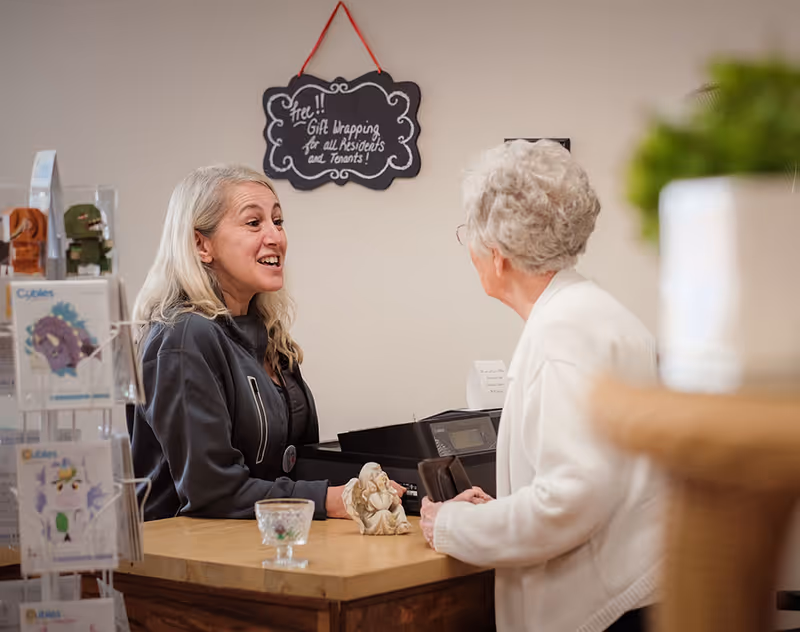 Two women engaged in conversation across a wooden counter inside a senior living facility. One woman with gray hair is seated behind the counter, smiling and talking, while the other elderly woman with white hair and glasses stands on the opposite side holding a wallet. A small decorative angel figurine and a glass bowl are on the counter. A sign hanging on the wall behind them reads 'Free!! Gift Wrapping for all Residents and Tenants!'