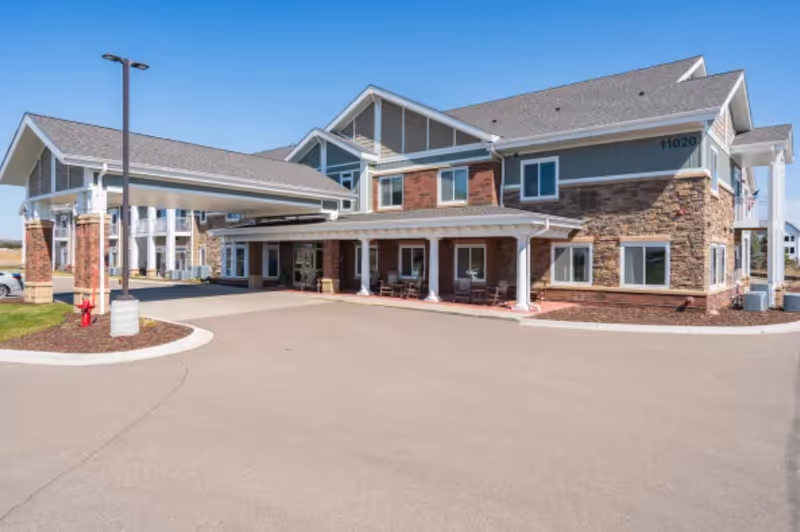 Exterior view of a two-story senior living facility building with a covered entrance, multiple windows, and a paved driveway under a clear blue sky.