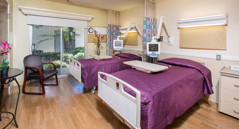 A bright and clean senior care facility bedroom with two hospital-style beds covered in purple bedspreads. Each bed has an adjustable overbed table and medical equipment mounted on the wall. There is a black chair with wooden armrests near a round table with a pink orchid on it. A large window with a view of greenery outside lets in natural light.