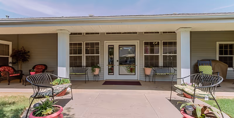 Front entrance of a building with a covered porch featuring white columns, black metal benches with cushions, potted plants, and outdoor seating. The building has gray siding and multiple windows around a central glass door.