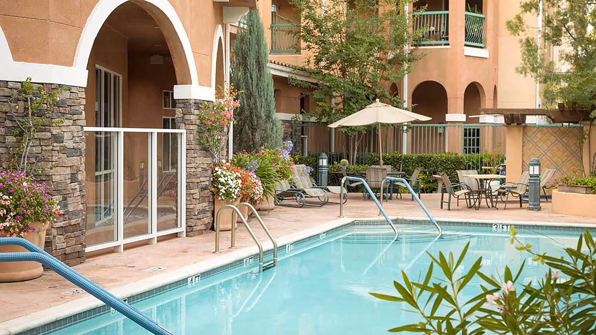 Outdoor swimming pool area at a residential facility with lounge chairs, tables with umbrellas, potted plants, and a building with arched doorways and balconies in the background.