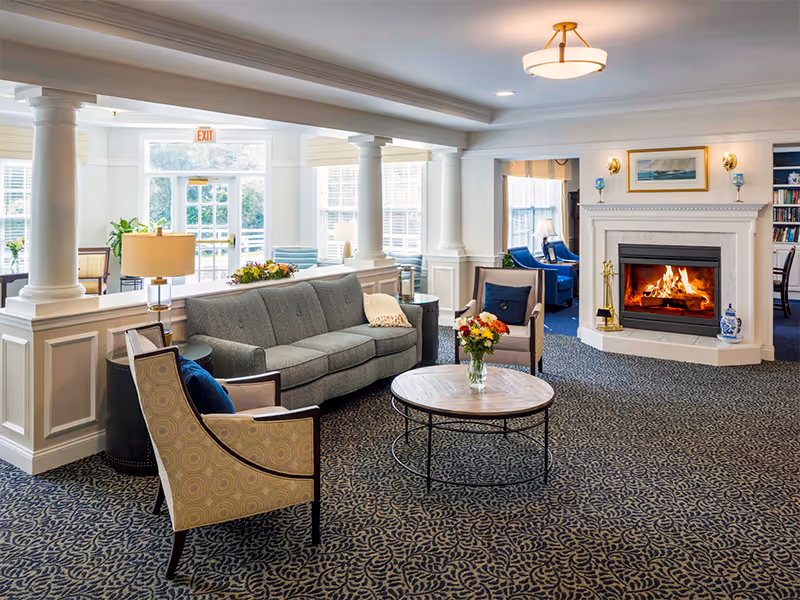 A cozy living room area with a gray sofa, two patterned armchairs, a round wooden coffee table with a vase of flowers, and a lit fireplace. The room has large windows letting in natural light, white columns, and a patterned carpet. There are additional seating areas visible in the background.