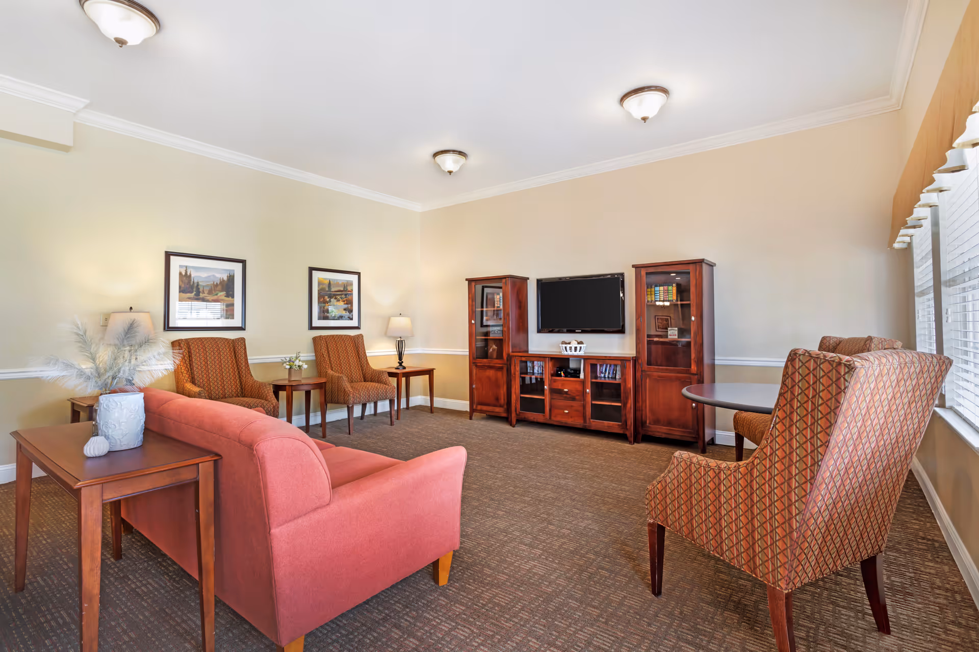A cozy living room with a red sofa, two patterned armchairs, two wooden side tables with lamps, a wooden entertainment center with a flat-screen TV, and two framed pictures on the wall. The room has beige walls, carpeted floor, and large windows with blinds letting in natural light.