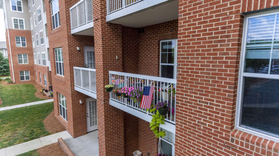 View of a brick apartment building with balconies decorated with potted flowers and an American flag. The building has multiple floors with white railings on the balconies and windows. There is a grassy area and paved walkways surrounding the building.