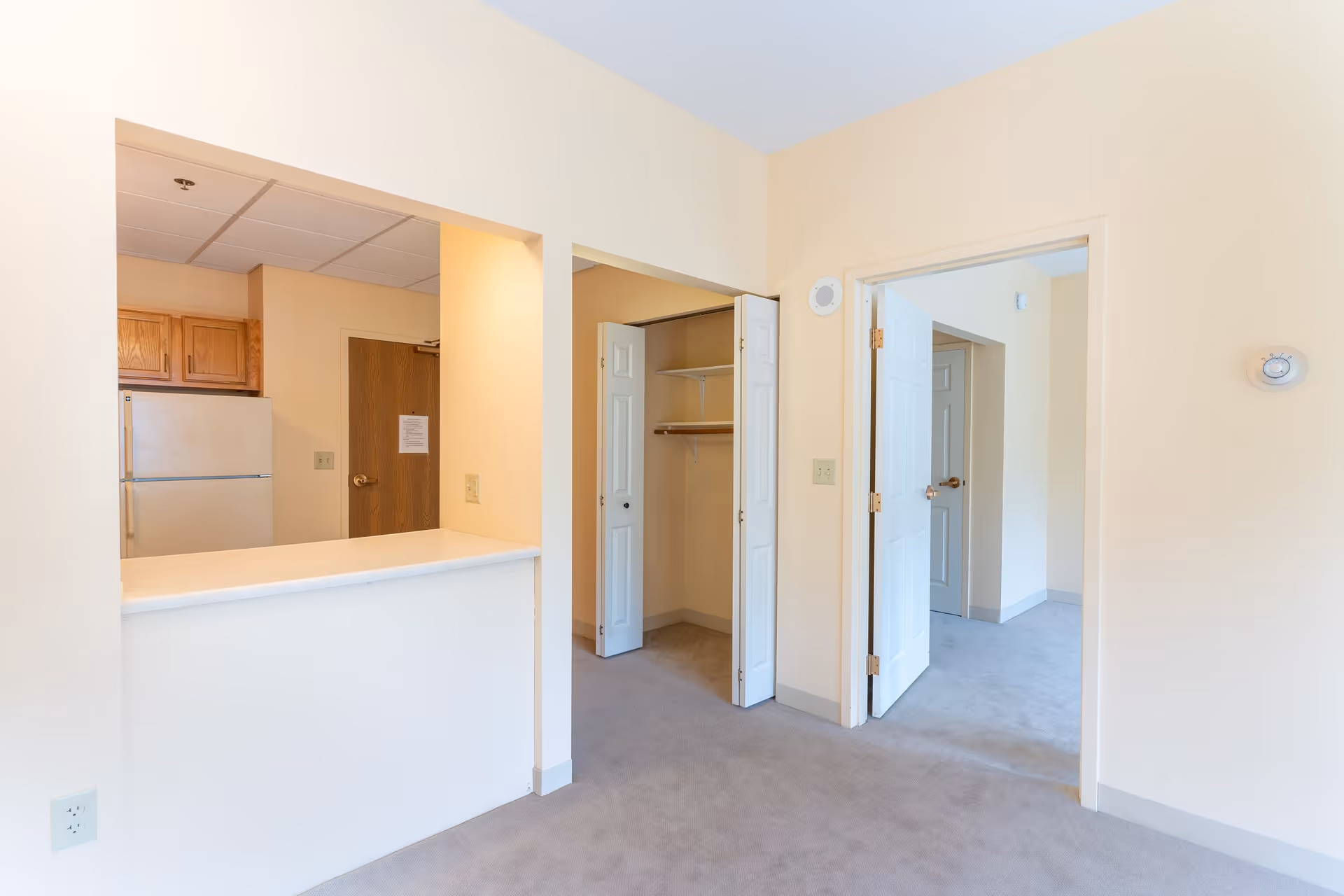 Interior view of an apartment unit showing a small kitchen area with a refrigerator and wooden cabinets, a countertop bar, an open closet with bi-fold doors, and an open doorway leading to another room. The walls are painted light beige and the floor is carpeted in gray.