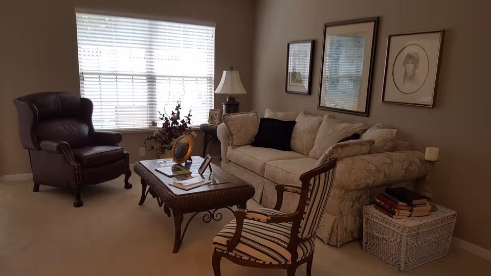 A cozy living room with a beige floral-patterned sofa adorned with cushions, a striped armchair, and a dark leather armchair. A wooden coffee table with glass top holds picture frames and books. A wicker side table with stacked books and a candle is next to the sofa. The room has beige walls with framed artwork and a large window with white blinds letting in natural light.