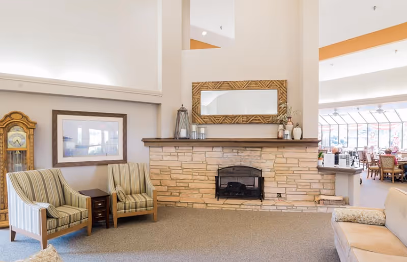 Spacious common room with a stone fireplace, decorative mirror, striped armchairs, a grandfather clock and a view into a dining area.