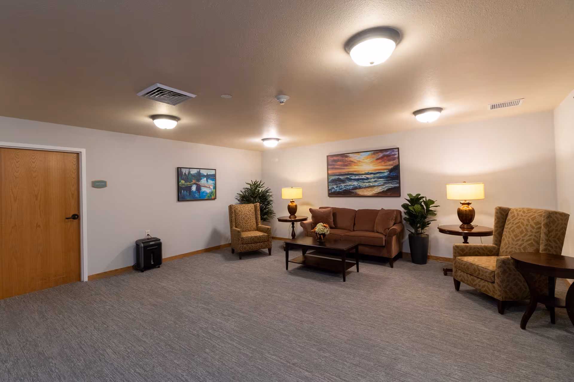 A cozy living room area with a brown sofa, two patterned armchairs, two side tables with lamps, a coffee table with a flower arrangement, two paintings on the walls, and two potted plants. The room has beige walls, a gray carpet, and ceiling lights.