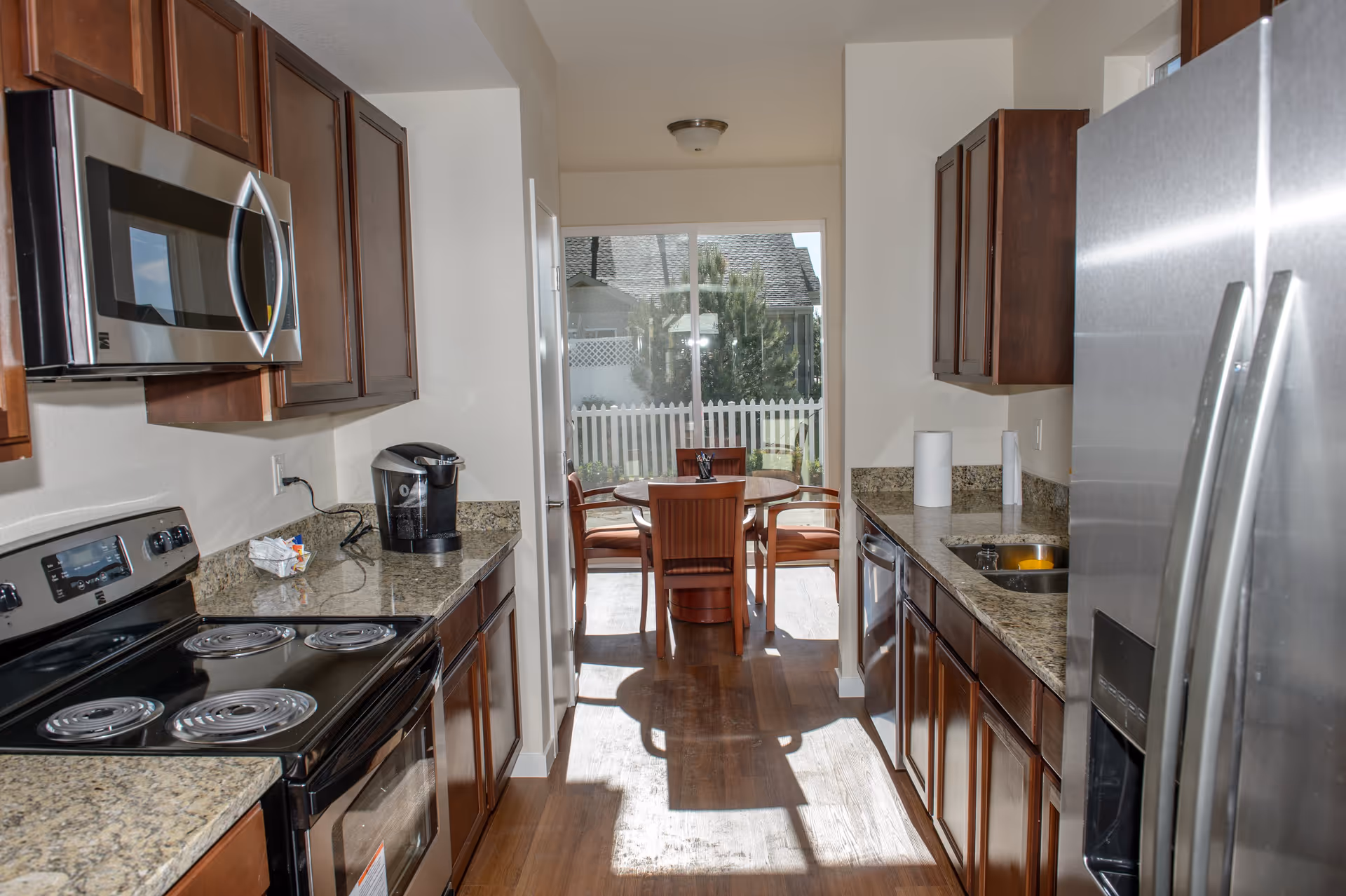 A modern kitchen with granite countertops, dark wooden cabinets, a stainless steel microwave, electric stove, dishwasher, and refrigerator. At the end of the kitchen, there is a round wooden dining table with four chairs, positioned in front of a large window with a view of a white picket fence and greenery outside.