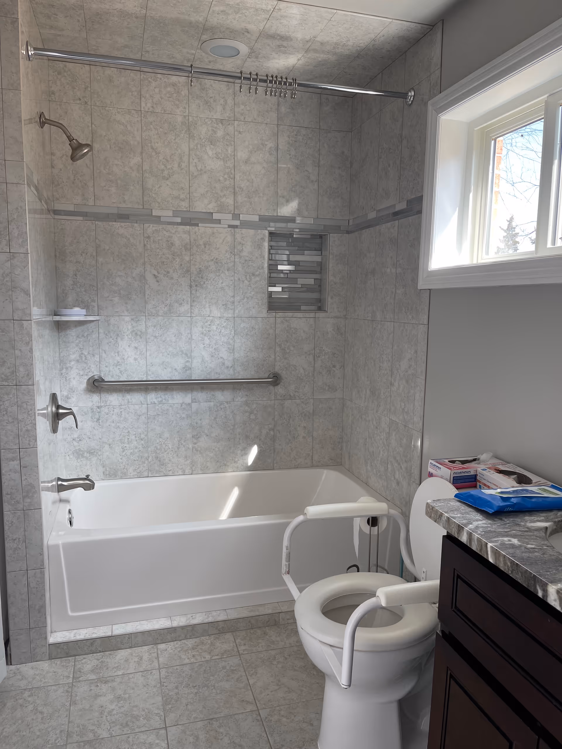 A bathroom with a bathtub and shower combination featuring gray tiled walls and a built-in niche with decorative tiles. There is a grab bar installed on the wall above the bathtub. A window lets in natural light. The toilet has white armrests for support, and a countertop with various items is visible next to it.