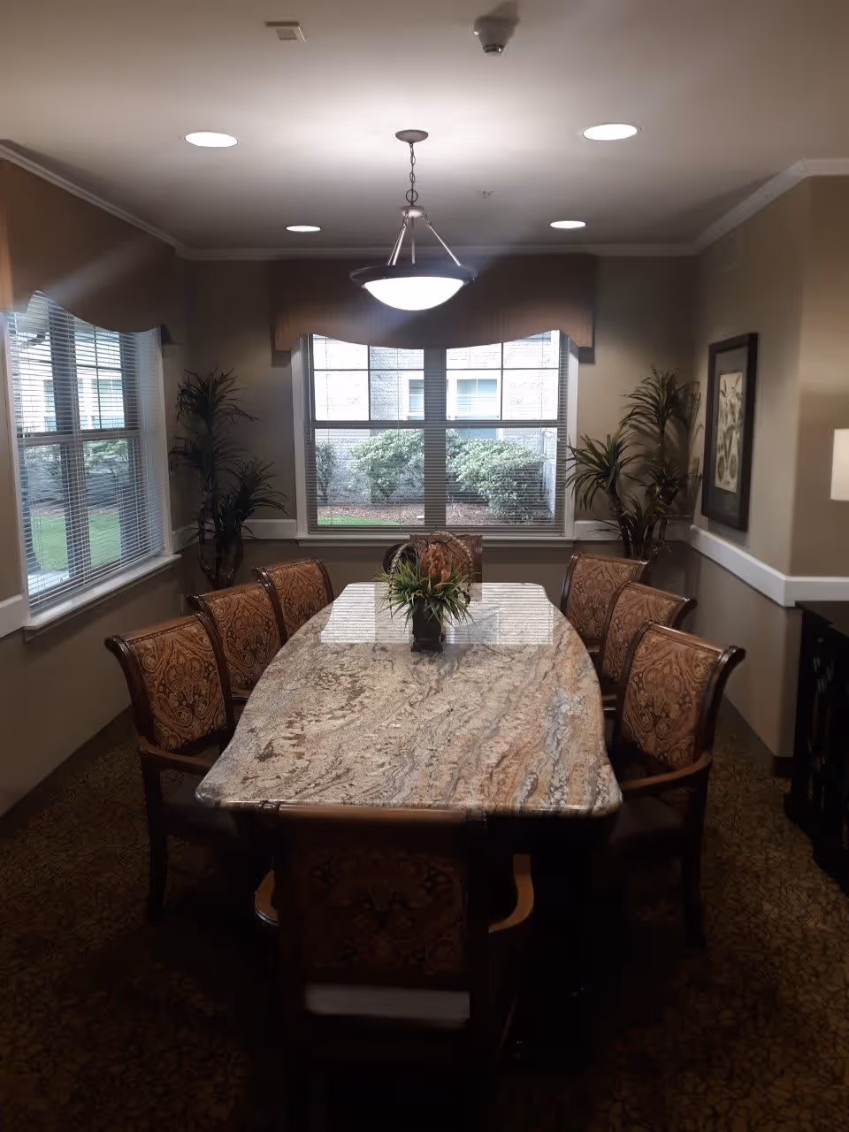 A dining room with a large rectangular marble table surrounded by eight ornate upholstered chairs. The room has beige walls, two large windows with blinds and valances, two potted plants in the corners, a hanging ceiling light fixture above the table, and a framed picture on the right wall.